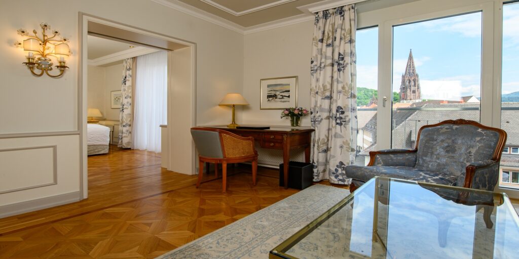Luxuriöse Hotel-Suite mit Parkett, eleganter Schreibtisch, gemütlicher Sitzgruppe und großem Fenster mit Kirchturmblick