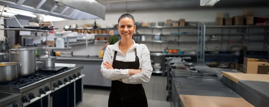 Köchin in professioneller Restaurantküche, lächelnd, mit verschränkten Armen, modernes Arbeitsumfeld, Edelstahlgeräte.