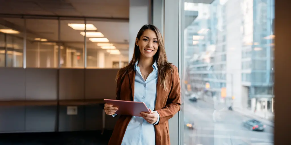 Eine lächelnde Frau im Büro hält ein Tablet, steht am Fenster mit Stadtblick im Hintergrund.