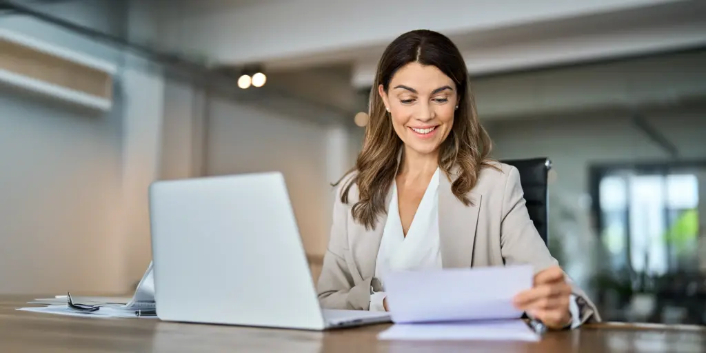 Frau arbeitet lächelnd am Laptop und liest Dokumente im Büro.