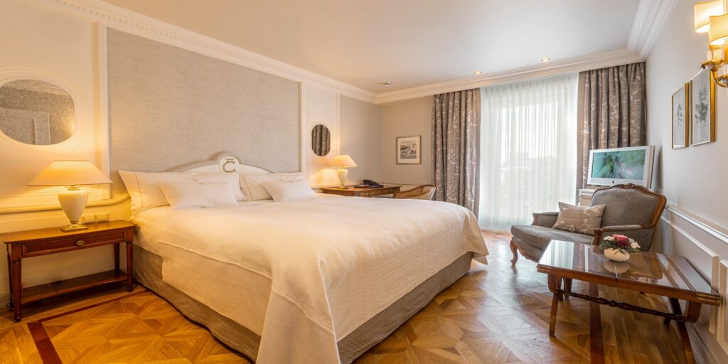Luxuriöses Hotelzimmer mit Kingsize-Bett, klassischem Parkett, gemütlichem Sitzbereich und hellem Balkonblick