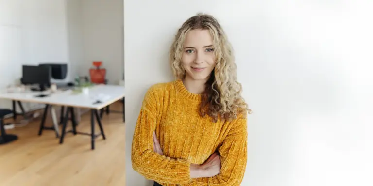 Eine lächelnde Frau in gelbem Pullover steht in modernem Büro mit gekreuzten Armen vor weißer Wand.