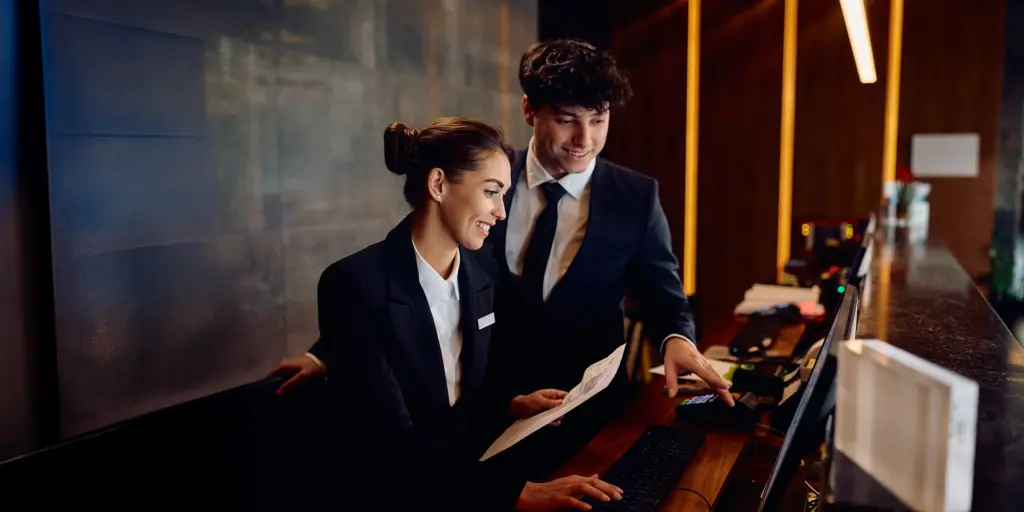 Zwei Hotelangestellte im Anzug arbeiten lächelnd an der Rezeption mit einem Computer und einem Dokument in der Hand.