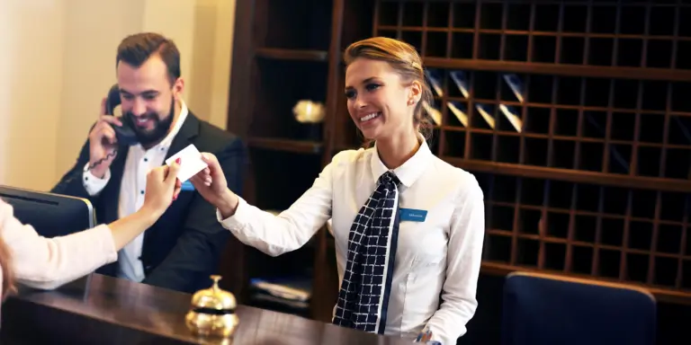 Hotelrezeptionistinnen überreichen eine Schlüsselkarte, während ein Kollege telefoniert. Empfangsbereich im Hintergrund.