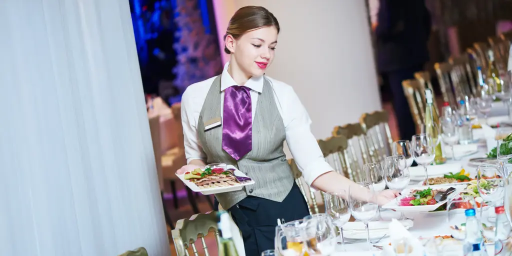 Kellnerin im Anzug serviert Essen bei einem festlichen Event, dekorierter Tisch mit Gläsern und Geschirr.