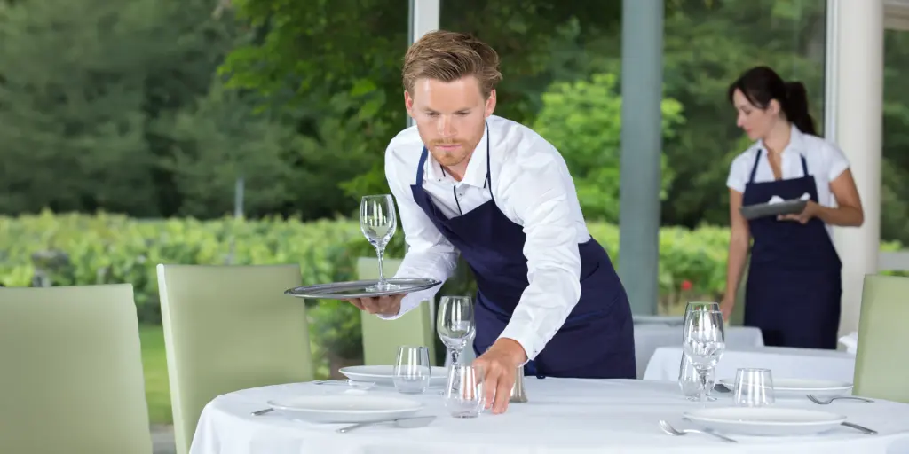 Ein Kellner deckt sorgfältig einen Tisch im Restaurant ein, die Szene ist von grünem Hintergrund umgeben.