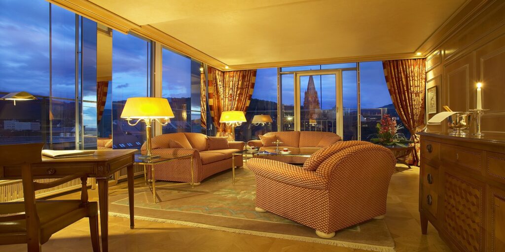 Luxuriöse Hotel-Suite mit Panoramafenstern, elegantem Wohnzimmer, warmem Licht und Blick auf Stadt und Kirche.