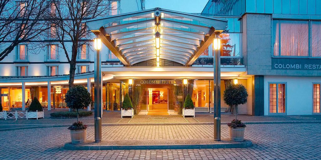 Luxuriöser Hoteleingang (Colombi Hotel) bei Abenddämmerung – beleuchtete Glasüberdachung, Pflanzkübel, Kopfsteinpflaster