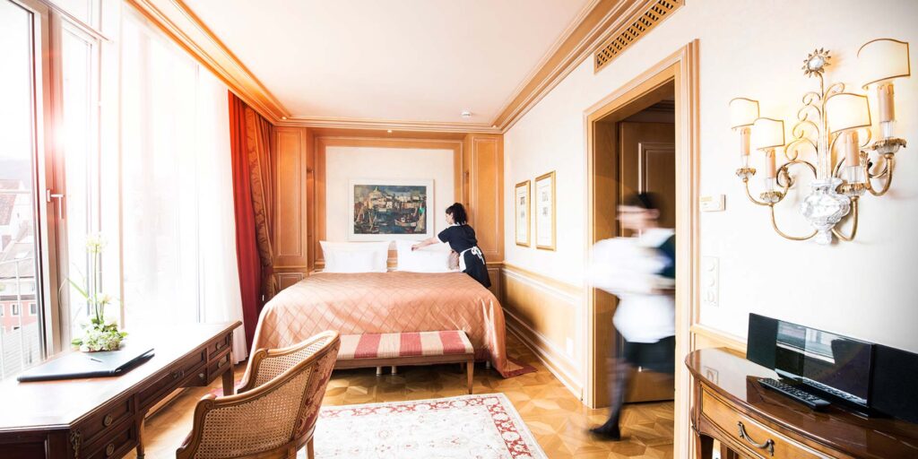 Luxushotelzimmer mit Doppelbett, klassischer Holz‑Inneneinrichtung, großem Fenster/Balkon und Servicepersonal.